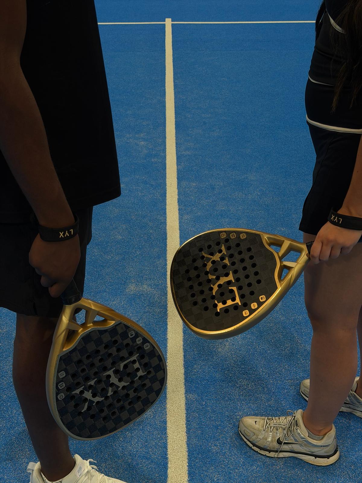 Two players facing off across a padel court centre line, both holding gold and black LYX Pro Range rackets
